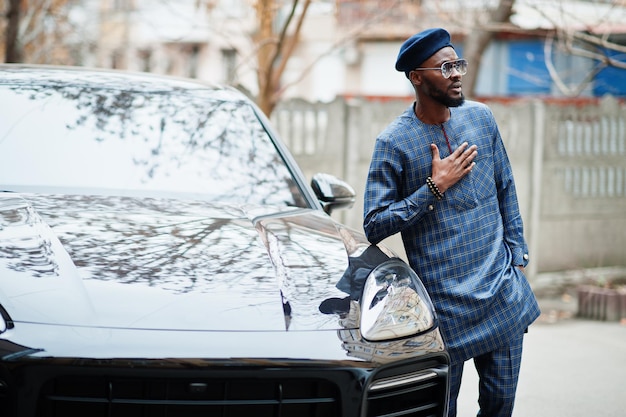 African guy in blue authentic costume, eyeglasses and beret. Fashionable nigerian man near black suv car.