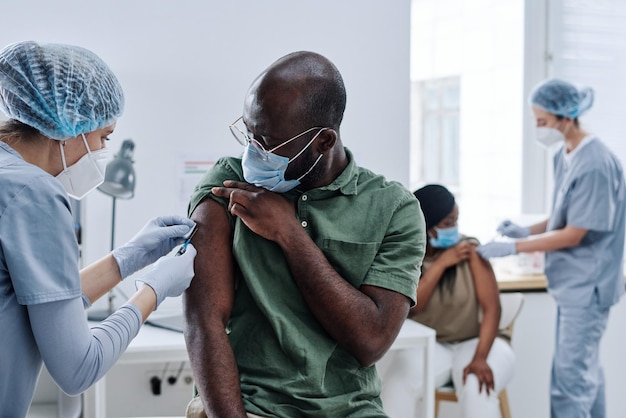 医者が病院への訪問中に注射をしている間、マスクをしたアフリカ人の男が肩を準備している