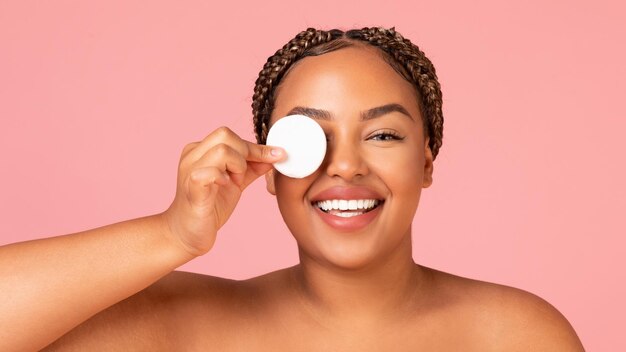 African lady covering eye with cotton pad over pink background