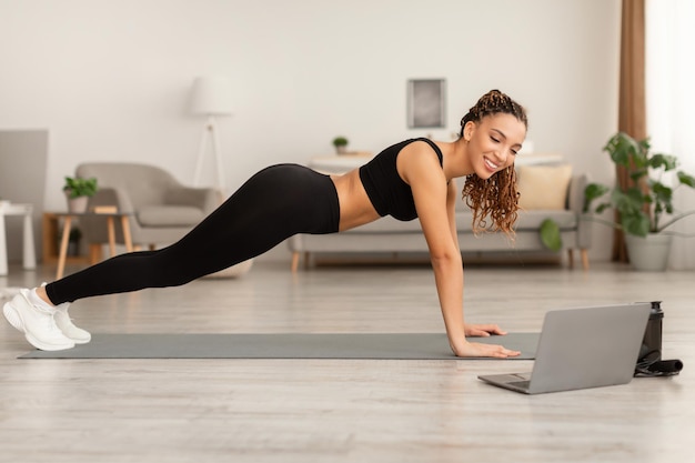 African lady near laptop standing in plank exercising at home