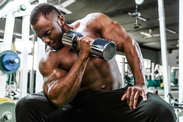 African male athlete doing exercises with dumbbells while sitting on a bench in the gym