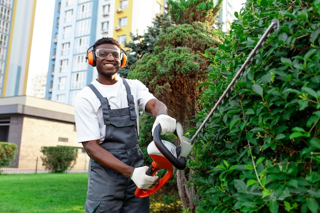 African male garden worker in uniform trims bushes with electric tool man works with brush cutter