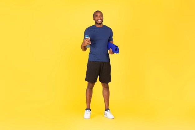 Photo african male holding water bottle and fitness mat yellow background