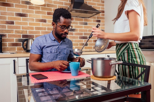 自宅でラップトップを使用しているアフリカの男性ヨーロッパの女性は、鍋のレンガの壁の炊飯器のフードの背景からプレートにスープを注いでいます