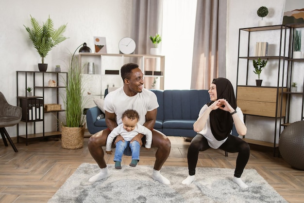 African Man holds son on his shoulders and muslim Woman in Workout Clothes are Doing Squat Exercises