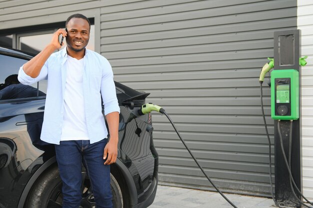 An african man is standing near an electric car waiting for it to charge at a charging station