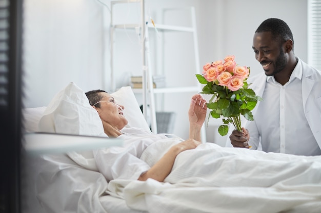 African man smiling and giving flowers to woman who lying on hospital bed at ward