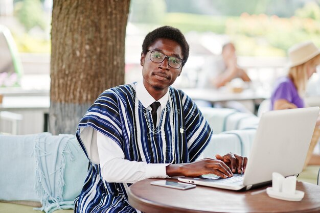 African man in traditional clothes and glasses sitting behind laptop at outdoor caffe and working