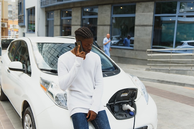 African man use smart phone while waiting and power supply connect to electric vehicles for charging the battery in car