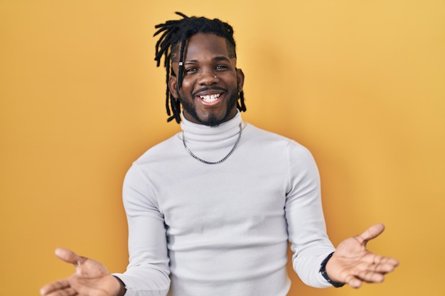 Photo african man with dreadlocks wearing turtleneck sweater over yellow background smiling cheerful with open arms as friendly welcome, positive and confident greetings