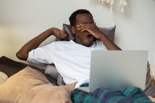 African man working lying on bed at home being tired yawning