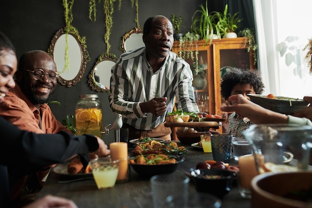 Foto uomo maturo africano che suggerisce il piatto per la sua famiglia mentre si siedono al tavolo nella sala da pranzo