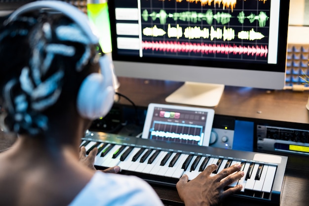 African or mixed-race young man touching keys of piano keyboard while sitting in front of computer screen and creating music