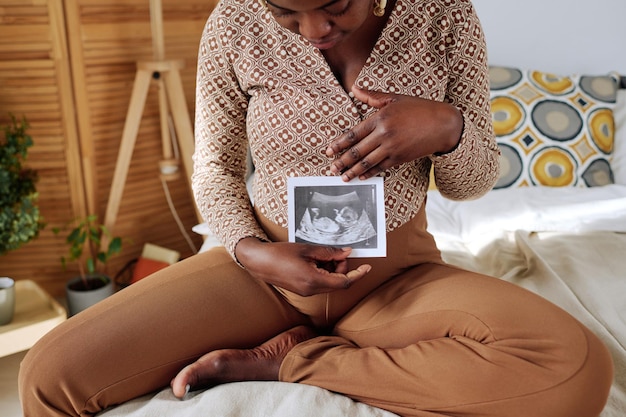 African pregnant girl looking at 3d ultrasound of her baby while sitting on bed in bedroom