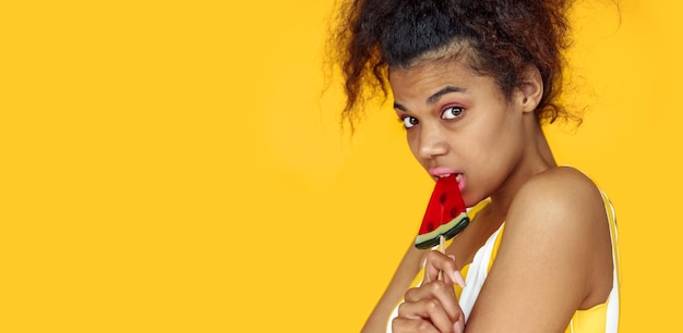 아프리카의 예쁜 십대 소녀가 노란색 여름 스튜디오 배경 배너에 사탕을 들고 있습니다.