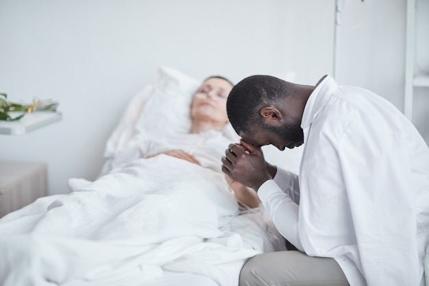 African sad man sitting in front of the hospital bed with his wife and worried about her health