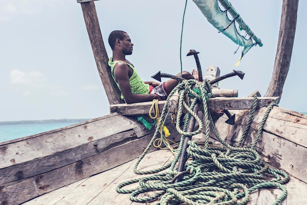 African sailor sitting on the bow of the boat next to a big\
anchor. zanzibar dhow in the sea, nungwi