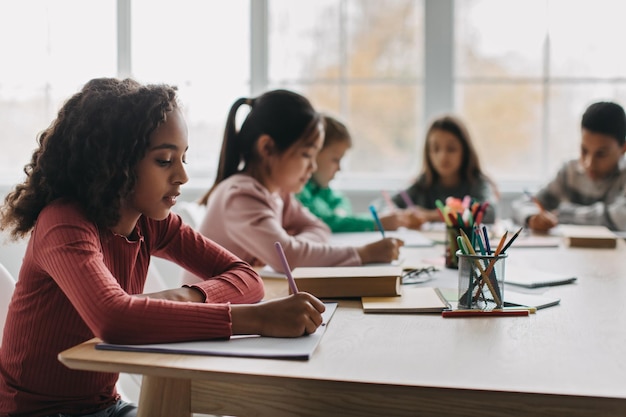 African Schoolgirl Taking Notes Sitting With Diverse Classmates In Classroom