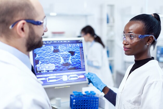 African scientist conducting chemistry experiment using computer with help from colleague. Multiethnic team of medical researchers working together in sterile lab wearing protection glasses and gloves