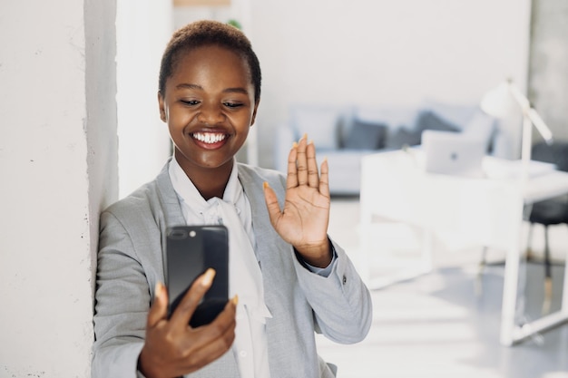 Donna d'affari sorridente africana che ha salutato la riunione virtuale sventolando la mano che tiene il video del telefono conver