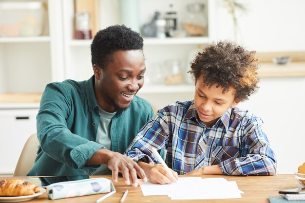 Photo african smiling father helping to his son doing his homework they sitting at the table in the room