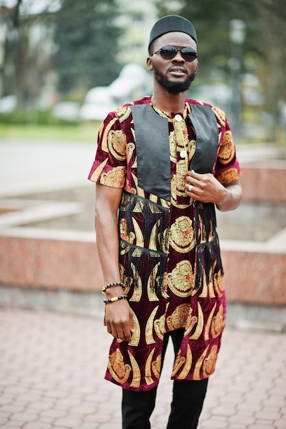 African stylish and handsome man in traditional outfit and black cap standing outdoor.