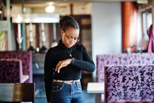 Африканская женщина в черном свитере позирует в кафе и смотрит на свои умные часы