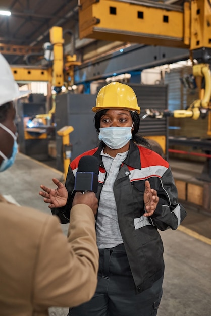 African woman in hardhat and uniform wearing protective mask talking with journalist during reportage in factory