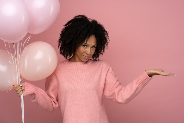 African woman holding a lot of balloons enjoys cool party wears pink sweater celebrates birthday