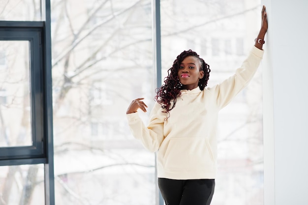 African woman in hoodie stand indoor apartment near large windows.