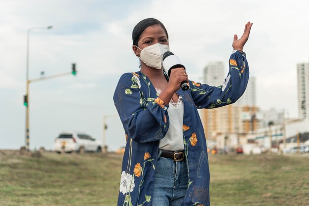 African woman journalist in face mask speaking into microphone while standing in city