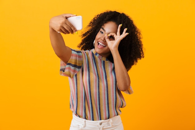 노란색 공간에 고립 된 포즈 아프리카 여자 괜찮아 제스처를 보여주는 휴대 전화로 selfie 걸릴.