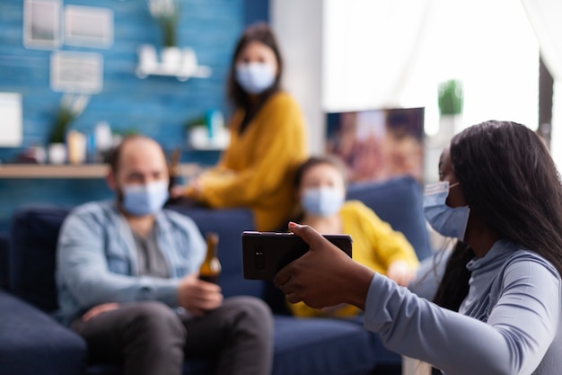 Photo african woman showing video clip to cheerful friends on smartphone in home living room wearing face mask as prevention against the spread and infection of covid19 while hanging out. conceptual image.