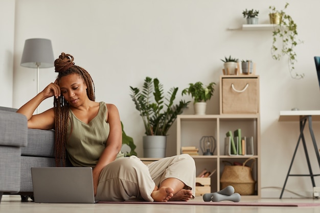 African woman sitting on the floor and watching sport blog on her laptop before sports training at home