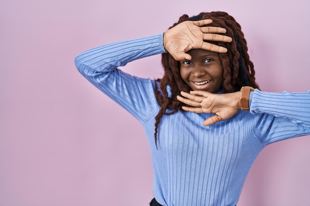 African woman standing over pink background smiling cheerful playing peek a boo with hands showing face. surprised and exited