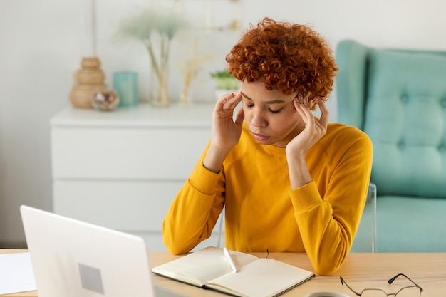 ストレスを感じているこめかみに触れるアフリカの女性 ホームオフィスの女の子 仕事にうんざりしている 頭痛 病気 こめかみをこする 額をこする 長いラップトップの使用 コンピュータビジョン症候群 オフィス症候群