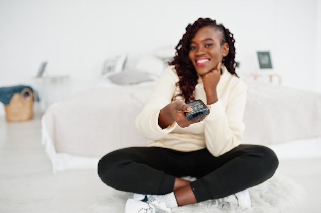 African woman watching tv at home and holding a remote control on bed.