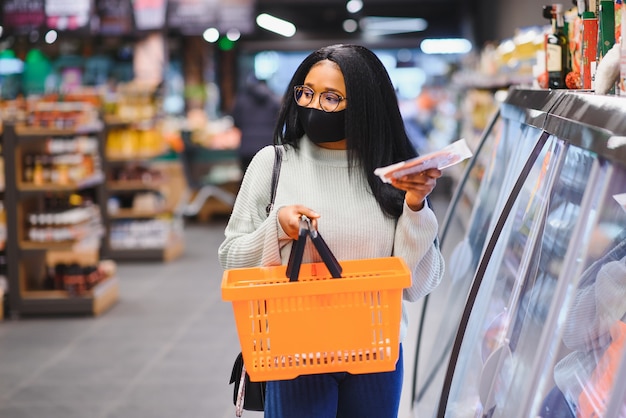 Donna africana che indossa una maschera medica usa e getta. fare la spesa al supermercato durante l'epidemia di pandemia di coronavirus. tempo di epidemia.