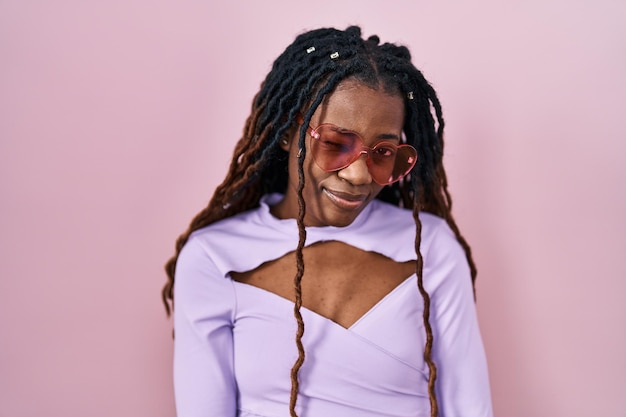 African woman with braided hair standing over pink background winking looking at the camera with sexy expression, cheerful and happy face.