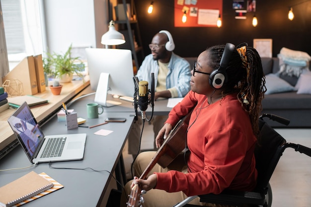 African woman with disability in headphones playing guitar while singing to the microphone during live broadcasting with music director in the background