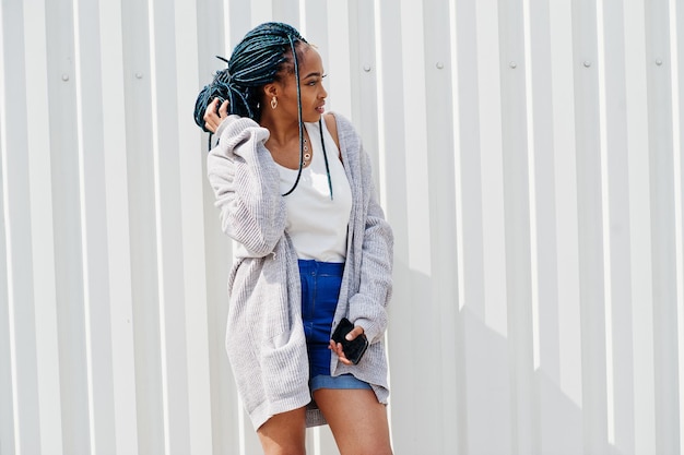 African woman with dreads hair in jeans shorts posed against white steel wall with mobile phone in hand