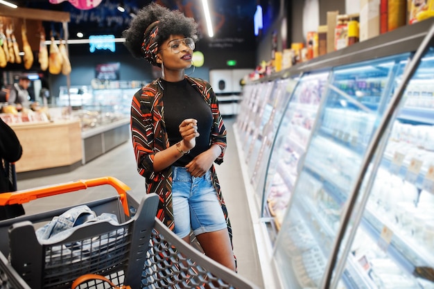 African woman with shopping cart choose yogurt bottle from fridge at supermarket