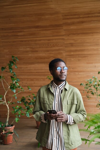 African young businessman in eyeglasses using mobile phone while standing at modern office