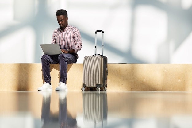 African young businessman sitting at the airport with luggage and working on laptop while waiting for his flight