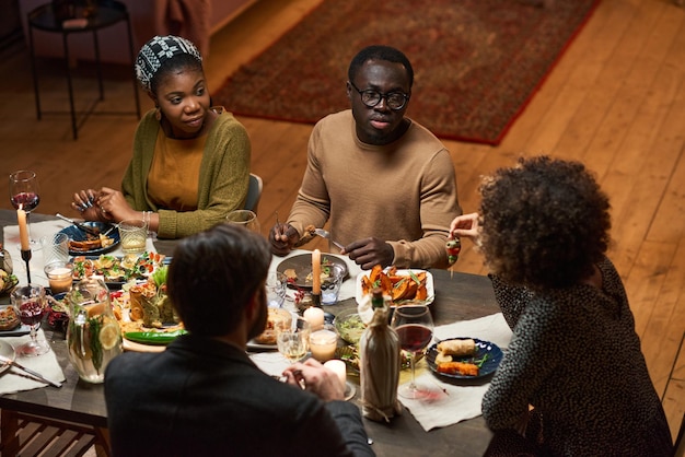 African young couple sitting at dining table and talking to their friends during dinner at home