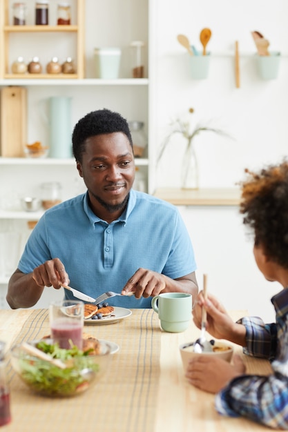 Il giovane padre africano fa colazione insieme a suo figlio al tavolo in cucina che sorridono e parlano