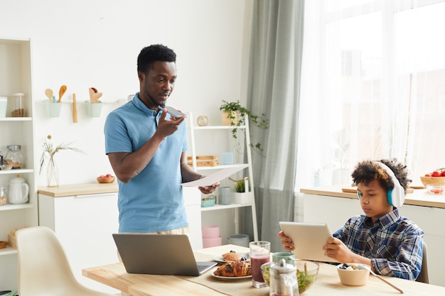 Giovane padre africano parlando al telefono ed esaminando i documenti che lavora a casa mentre suo figlio gioca sulla tavoletta digitale in cucina