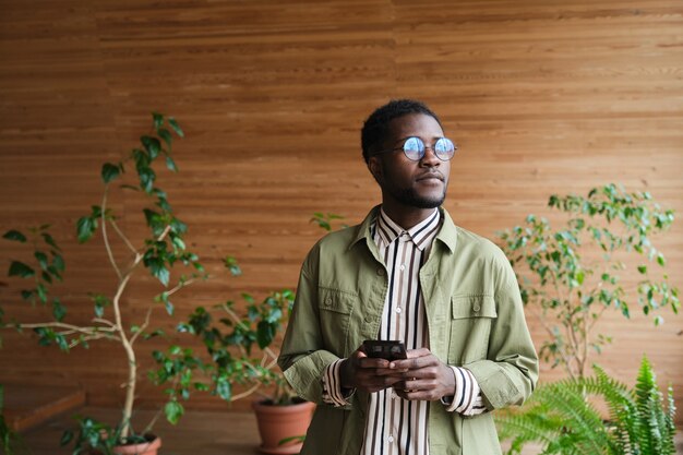 African young man in eyeglasses using mobile phone while standing in modern hall