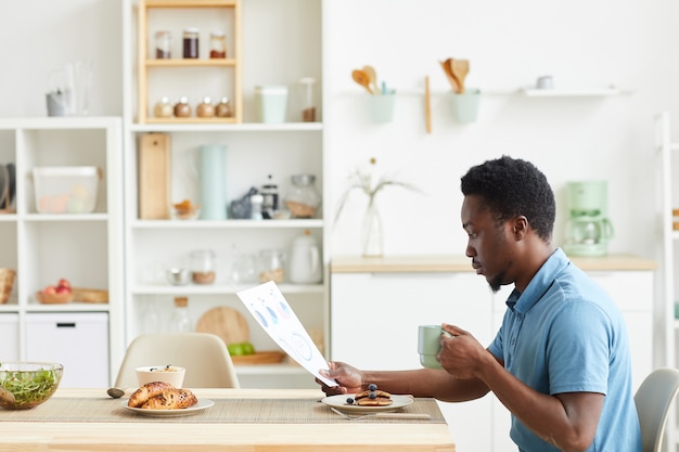 彼は朝食と読書のビジネス契約を持っているキッチンのテーブルに座っているアフリカの若い男