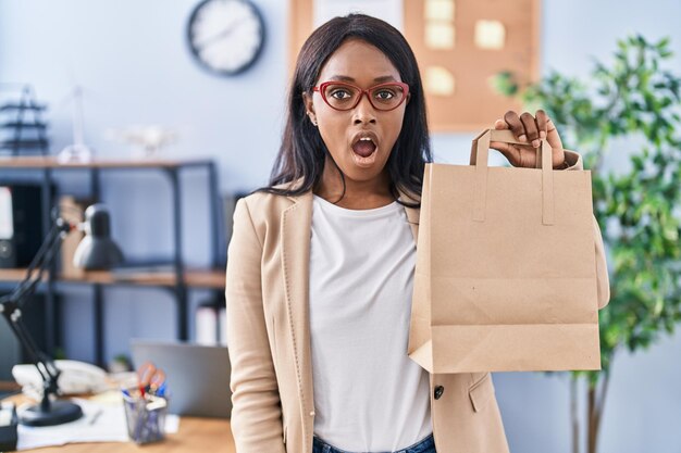 African young woman holding take away bag at the office scared and amazed with open mouth for surprise disbelief face